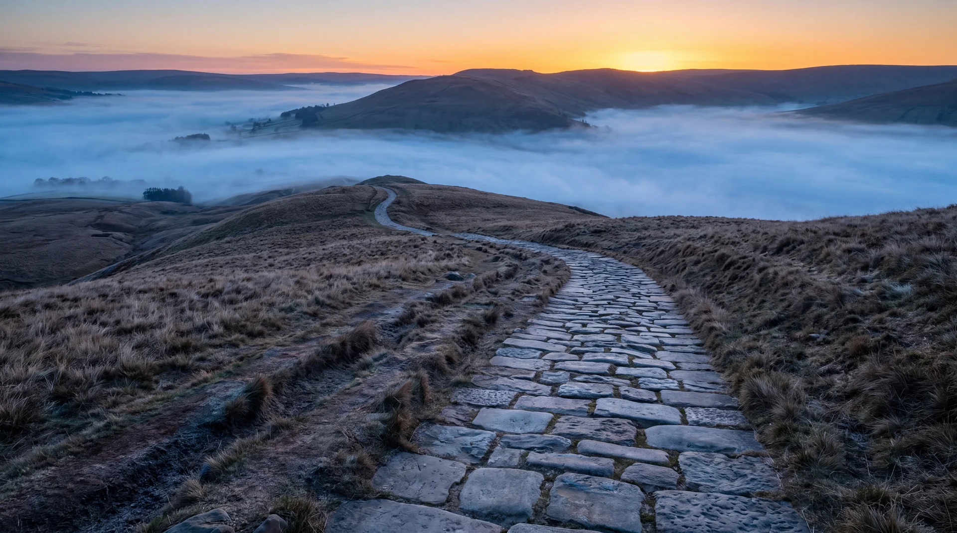 An ancient worn stone road climbing through open hills toward a sunrise, the paving smoothed by long use, the way clearly visible and open ahead, cold blue valley mist below and warm gold sky at the crest
